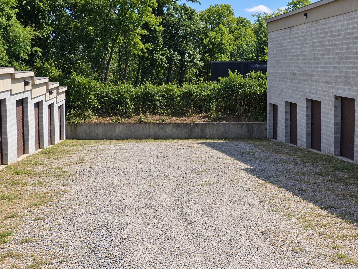 The wide gravel drive aisle between two block storage buildings at Safe Haven Storage, with brown roll-up doors down both sides and a hedge and trees at the far end